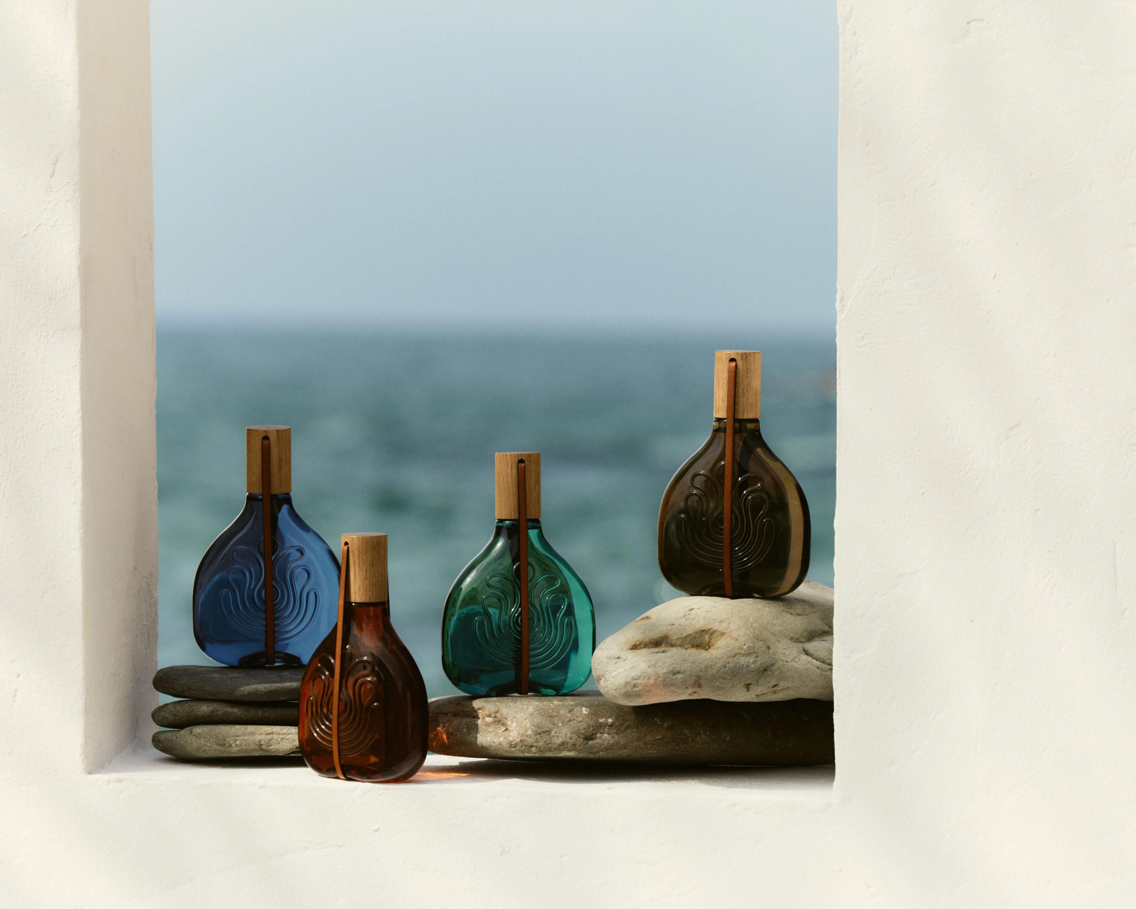 The four Puig absolute colognes displayed in colored glass bottles on a windowsill with the sea in the background.